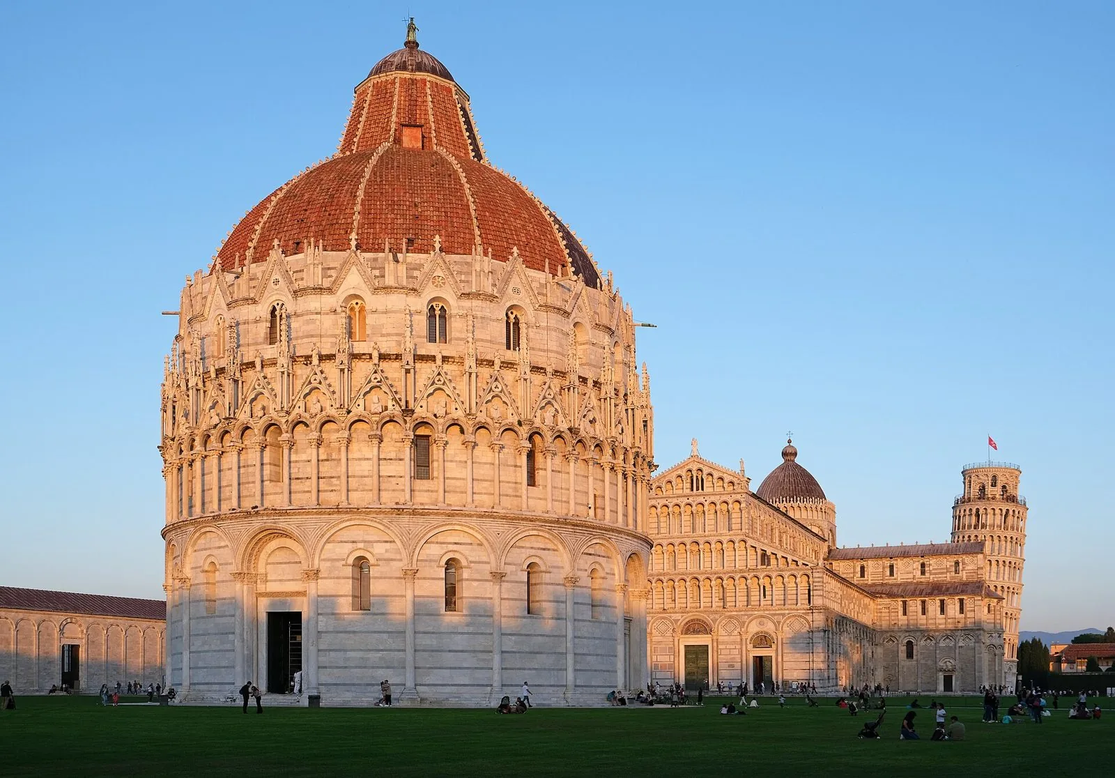Il Battistero di Pisa illuminato dal sole del tramonto, con il Duomo e la Torre pendente sullo sfondo e il prato di Piazza dei Miracoli