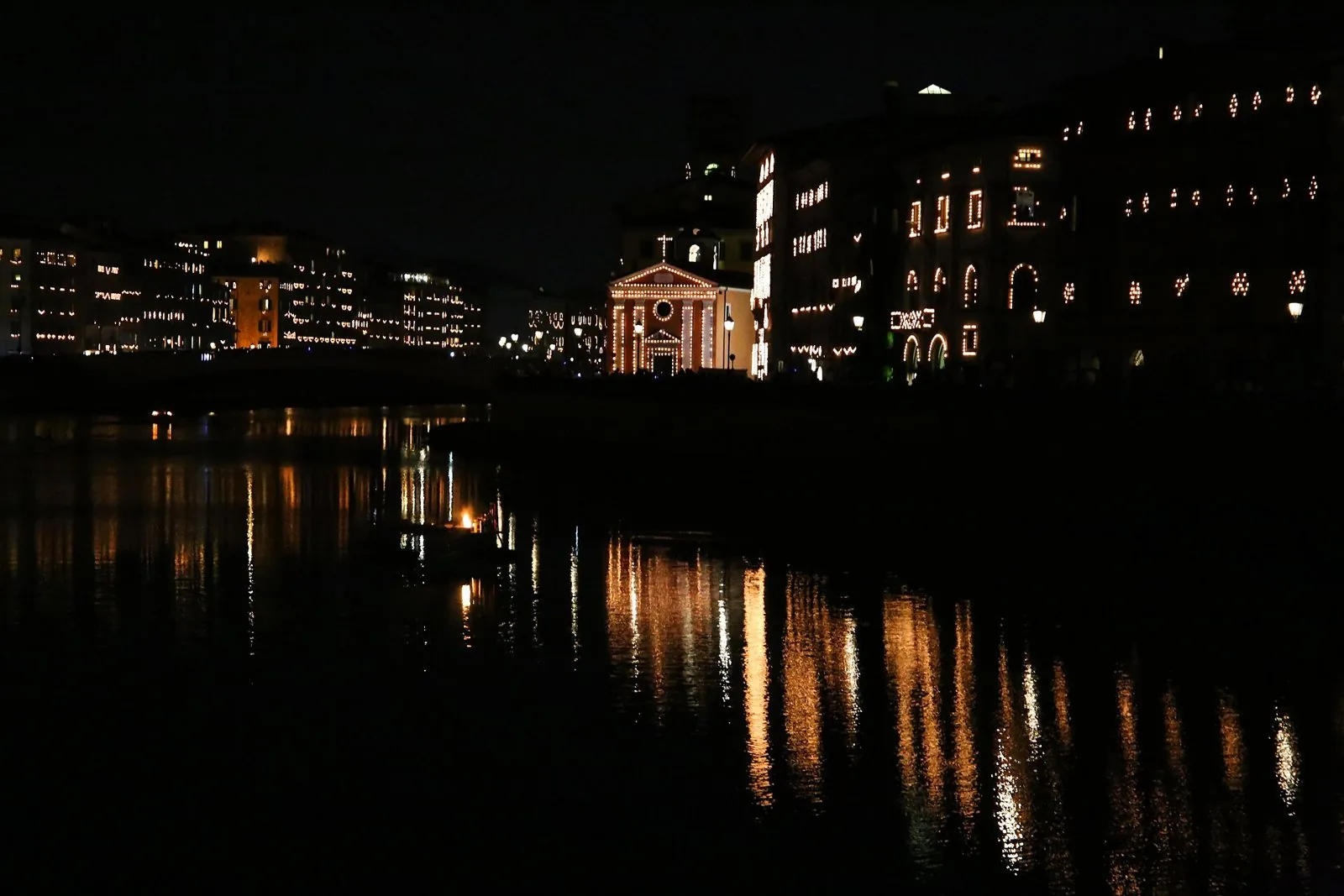 Lungarno di Pisa di notte durante la Luminara, con facciate di palazzi e di una chiesa disegnate dai lumini e riflesse nell'Arno