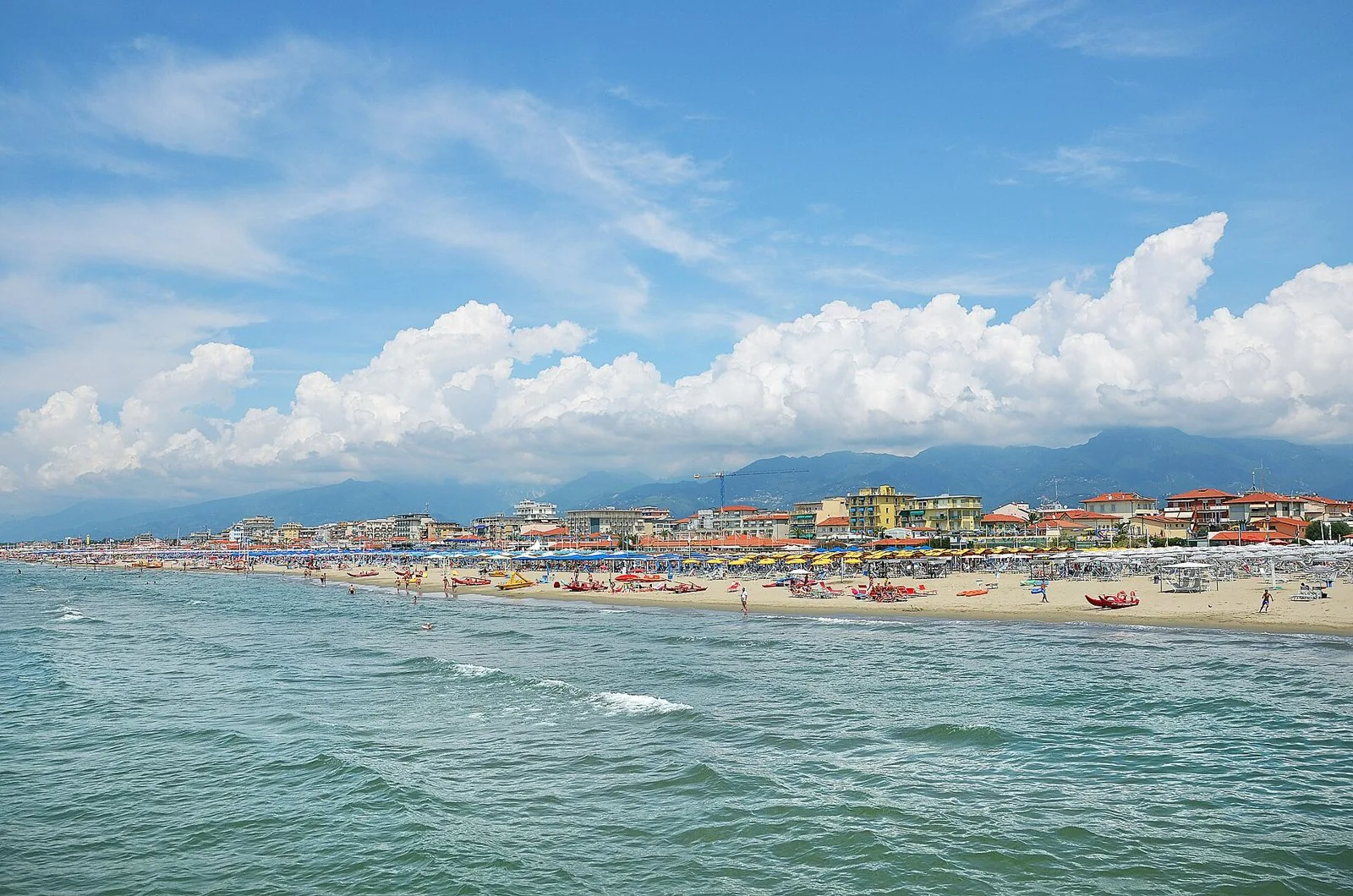 La spiaggia di Lido di Camaiore vista dal pontile, con ombrelloni colorati, palazzine sul lungomare e le Alpi Apuane tra le nuvole